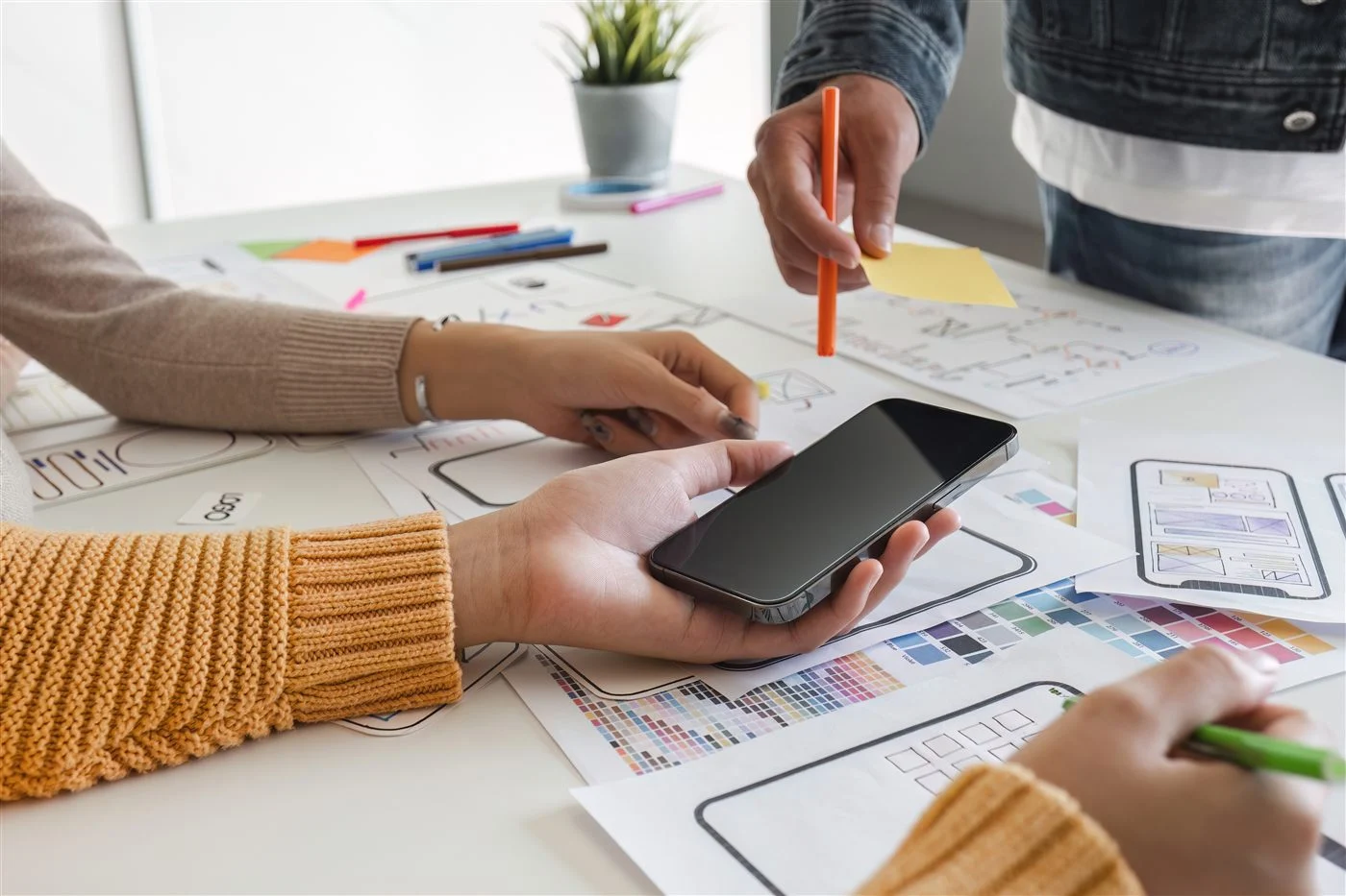 Des mains tiennent un téléphone au-dessus de maquettes d’écrans dessinées sur papier